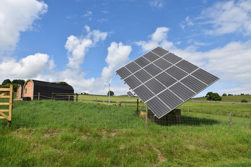 Solar panel array in a grassy field near wooden glamping pods under a partly cloudy sky.