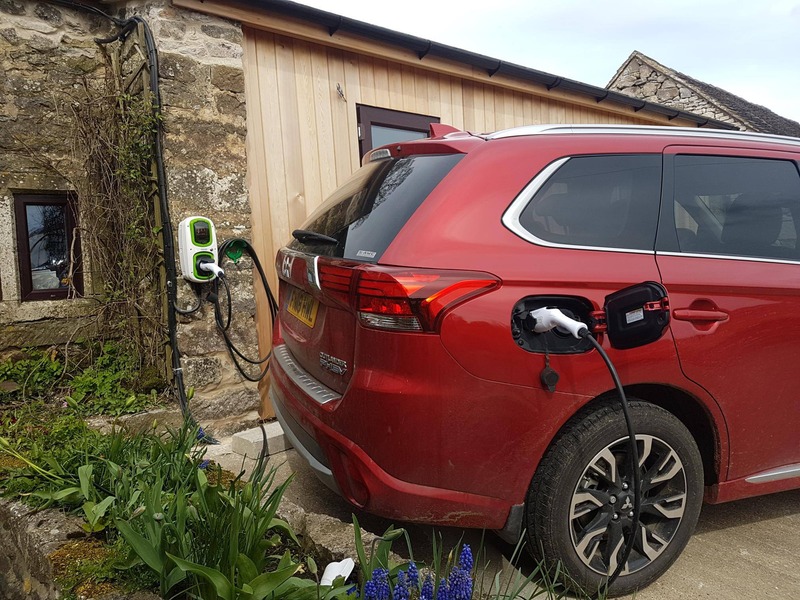 Red electric SUV charging at a wall-mounted EV charger beside a stone and timber cottage.