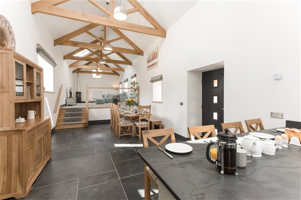Open plan living area with slate topped breakfast bar in foreground, leading to the oak dinning table, slate floor, exposed oak beams and lounge area up 4 steps with grey carpet and green/blue sofas and chairs