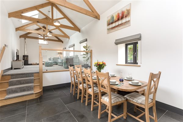 Open plan living area with slate floored dinning room in foreground, 4 steps leaving to lounge area. Glass balustrade, exposed oak beams, black wood burner.