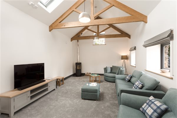 Lounge area with grey carpet, large TV, exposed oak beams, black wood burner and green/blue sofas and swivel chair
