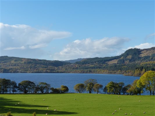 a field and a blue loch in scotland, with hills in the distance 