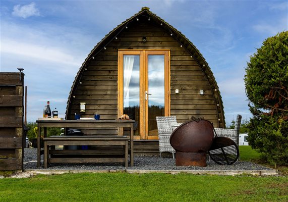Exterior of a cosy wooden glamping pod at Galloway Retreats in Dumfries and Galloway, Scotland, featuring glass double doors, outdoor seating, a fire pit and a peaceful countryside setting.