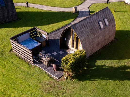 Aerial view of a luxury glamping pod surrounded by green countryside, featuring a private outdoor hot tub, enclosed patio and seating area. Aerial view of a luxury glamping pod surrounded by green countryside, featuring a private outdoor hot tub, enclosed patio and seating area.
