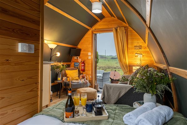 Romantic luxury glamping pod bedroom with a cosy double bed, warm wooden interiors, soft lighting and a breakfast tray, overlooking beautiful countryside views. Romantic luxury glamping pod bedroom with a cosy double bed, warm wooden interiors, soft lighting and a breakfast tray, overlooking beautiful countryside views.