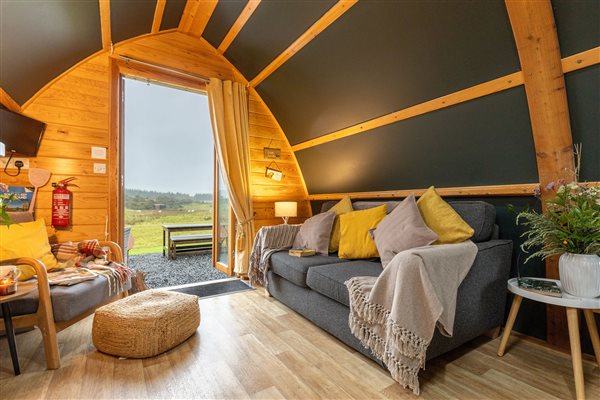 Cosy luxury glamping pod interior at Galloway Retreats in Dumfries and Galloway, Scotland, featuring a comfortable sofa, warm wooden beams, stylish furnishings and countryside views through the glass doors.