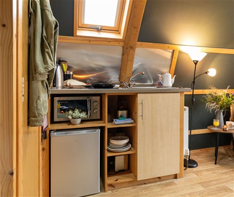 Compact kitchenette inside a luxury glamping pod at Galloway Retreats, featuring a mini fridge, microwave, sink, storage cupboards, wooden furnishings and warm lighting.