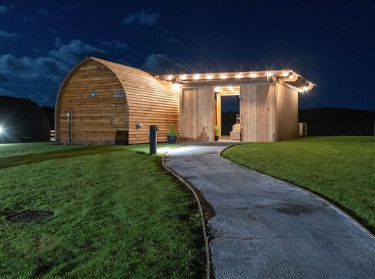 Luxury wooden glamping pod illuminated at night, with a curved pathway leading to the entrance, outdoor lighting and surrounding green lawns. Luxury wooden glamping pod illuminated at night, with a curved pathway leading to the entrance, outdoor lighting and surrounding green lawns.