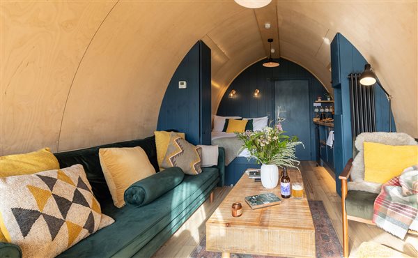 Stylish luxury glamping pod interior featuring a cosy lounge with green velvet sofa, comfortable armchair, decorative cushions and wooden coffee table leading towards the kitchenette. Stylish luxury glamping pod interior featuring a cosy lounge with green velvet sofa, comfortable armchair, decorative cushions and wooden coffee table leading towards the kitchenette.