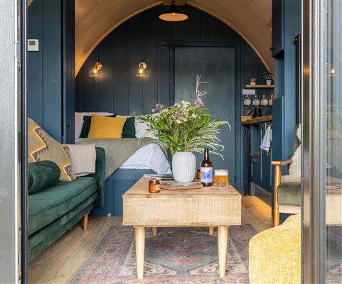 Stylish interior of a premium luxury glamping pod at Galloway Retreats, featuring a cosy double bed, comfortable green sofa, wooden coffee table, elegant dark walls and warm ambient lighting.