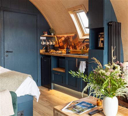 Cosy luxury glamping pod interior featuring a modern kitchenette with dark blue cabinets, wooden worktops, skylight window and stylish furnishings.