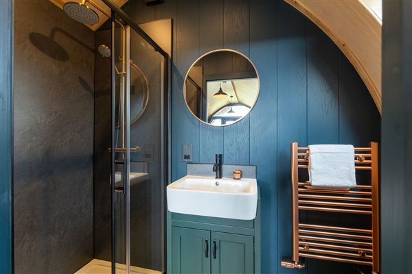 Modern bathroom at Galloway Retreats luxury glamping pod, featuring a walk-in shower, stylish blue walls, vanity sink, round mirror and wooden towel rail. Modern bathroom at Galloway Retreats luxury glamping pod, featuring a walk-in shower, stylish blue walls, vanity sink, round mirror and wooden towel rail.