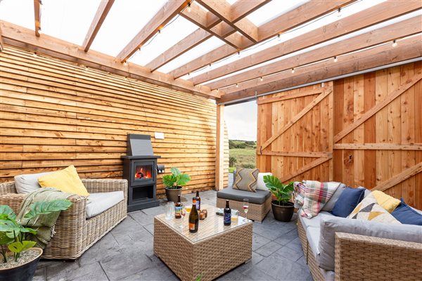 Luxury glamping pod’s covered outdoor lounge featuring comfortable rattan sofas, cosy cushions, a coffee table, wood-burning stove and wooden pergola overlooking the countryside.