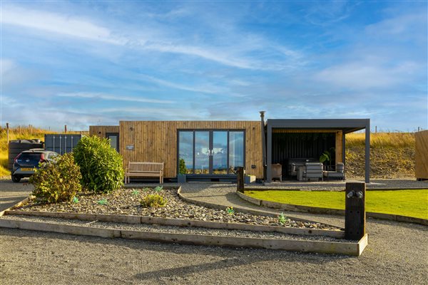 Large luxury countryside cabin with modern timber exterior, floor-to-ceiling glass doors, covered outdoor seating area, landscaped gardens and spacious private driveway.
