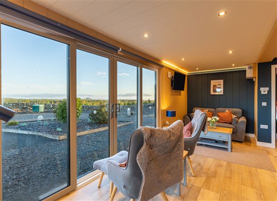 Spacious luxury countryside cabin living room with comfortable armchairs, stylish modern furnishings, floor-to-ceiling glass doors and beautiful views of the private outdoor terrace and surrounding hills.