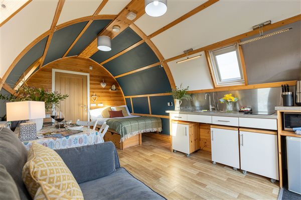 Cosy luxury glamping pod interior featuring a modern kitchenette, dining area, comfortable sofa and beautiful curved wooden ceiling. Cosy luxury glamping pod interior featuring a modern kitchenette, dining area, comfortable sofa and beautiful curved wooden ceiling.