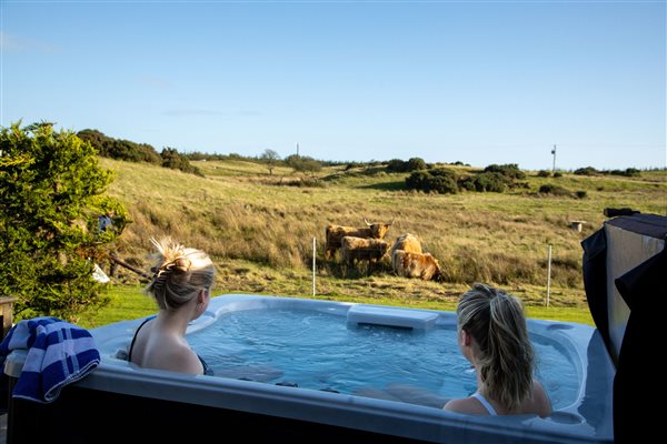 Two guests relaxing in a private outdoor hot tub at a luxury glamping pod, enjoying beautiful countryside views with highland cows grazing in the nearby fields. Two guests relaxing in a private outdoor hot tub at a luxury glamping pod, enjoying beautiful countryside views with highland cows grazing in the nearby fields.