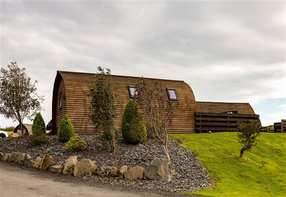 Exterior of a luxury wooden glamping pod surrounded by landscaped gardens, decorative shrubs, natural stone borders and green lawns in a peaceful countryside setting.