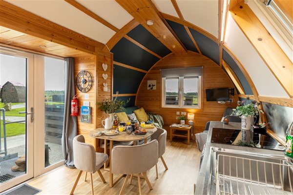 Bright open-plan living and dining area inside a luxury glamping pod, featuring exposed wooden beams, a dining table, comfortable seating and patio doors overlooking the countryside. Bright open-plan living and dining area inside a luxury glamping pod, featuring exposed wooden beams, a dining table, comfortable seating and patio doors overlooking the countryside.
