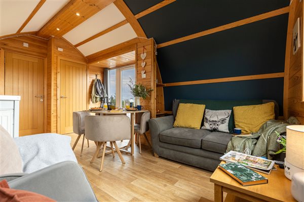 Cosy luxury glamping cabin living area with comfortable grey sofa, yellow cushions, wooden beams, stylish dining table and warm natural wood interiors.