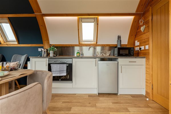 Bright, modern kitchen inside a luxury glamping cabin, featuring white fitted cupboards, worktops, cooking facilities, a sink and warm natural wooden beams.