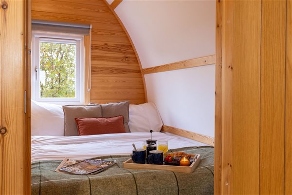 Cosy bedroom inside a luxury wooden glamping pod, featuring a comfortable double bed, soft cushions, natural wood panelling and a breakfast tray. Cosy bedroom inside a luxury wooden glamping pod, featuring a comfortable double bed, soft cushions, natural wood panelling and a breakfast tray.