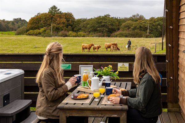 Two guests enjoying breakfast on the private outdoor decking of a luxury glamping pod, overlooking horses grazing in peaceful green countryside. Two guests enjoying breakfast on the private outdoor decking of a luxury glamping pod, overlooking horses grazing in peaceful green countryside.