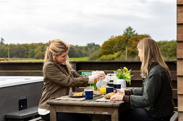 Two guests enjoying breakfast together on the private outdoor decking of a luxury glamping pod with hot tub, overlooking peaceful countryside views.