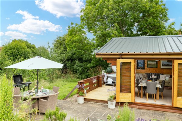 Log cabin with open double doors looking into the dining area, outdoor seating and veranda with hot tub